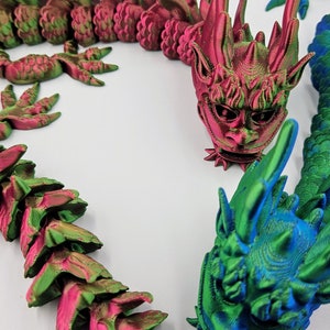May include: 3D-printed articulated dragon in shades of green, pink, and blue. The dragon features a detailed head and body segments, with a textured surface. The design includes sharp spines and a stylized face, creating a fantasy creature.