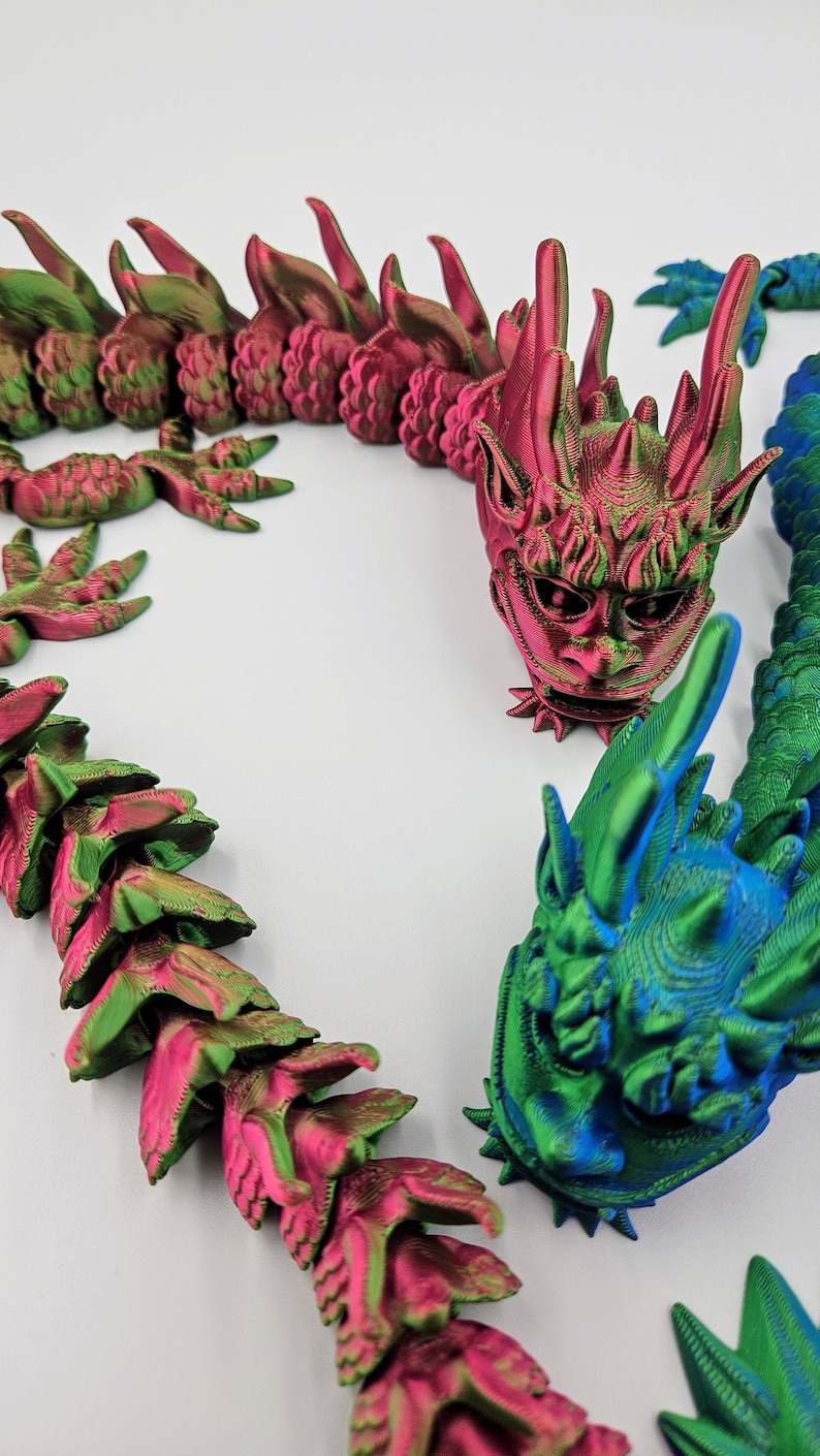 May include: 3D-printed articulated dragon in shades of green, pink, and blue. The dragon features a detailed head and body segments, with a textured surface. The design includes sharp spines and a stylized face, creating a fantasy creature.