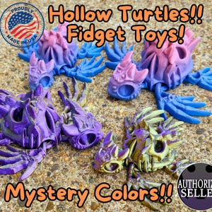 Peut inclure: Quatre jouets de stimulation en forme de tortues creuses, de couleur violette, bleue et dor&eacute;e. Les jouets sont fabriqu&eacute;s aux &Eacute;tats-Unis et sont disponibles en couleurs myst&egrave;re. L'image comprend &eacute;galement le texte "Hollow Turtles!! Fidget Toys!" et "Mystery Colors!!"