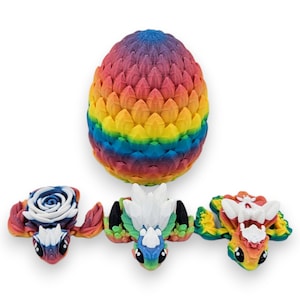 Puede incluir: Tres peque&ntilde;as figuras de drag&oacute;n de colores, cada una con un dise&ntilde;o diferente en la espalda, est&aacute;n sentadas frente a un huevo de drag&oacute;n m&aacute;s grande, de color arco&iacute;ris.