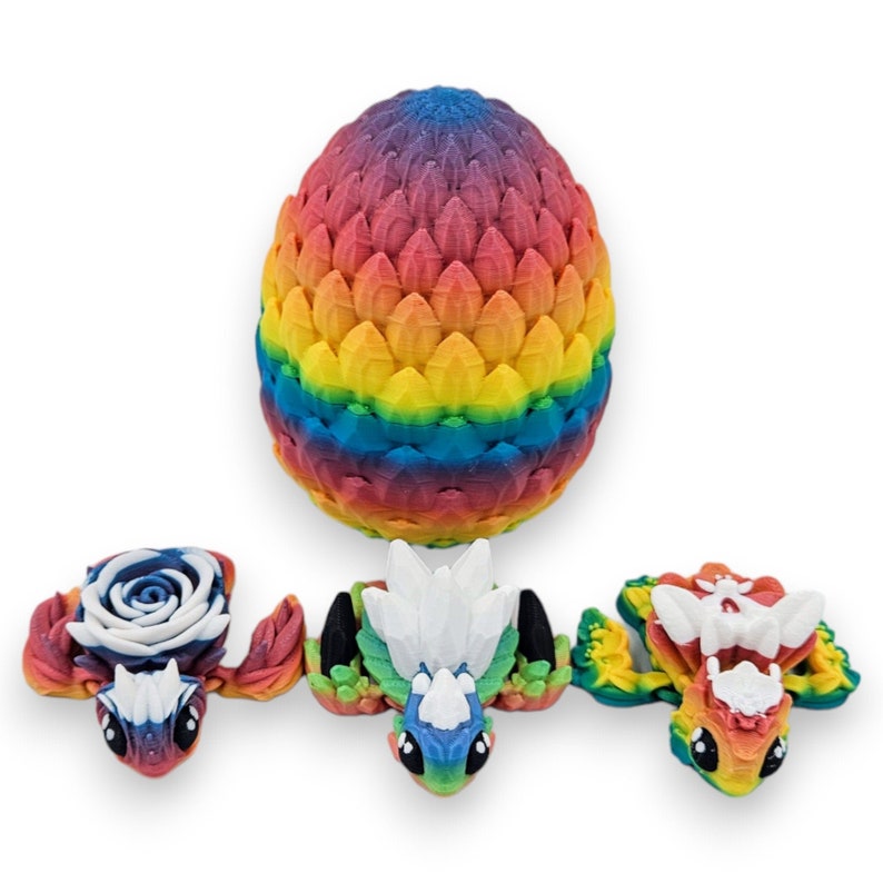 Puede incluir: Tres peque&ntilde;as figuras de drag&oacute;n de colores, cada una con un dise&ntilde;o diferente en la espalda, est&aacute;n sentadas frente a un huevo de drag&oacute;n m&aacute;s grande, de color arco&iacute;ris.