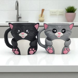 May include: Two cat-shaped mugs. One is black, the other gray. Both have white bellies, pink ears, noses, and paw prints. The mugs have large eyes and handles. The mugs are sitting on a white surface.