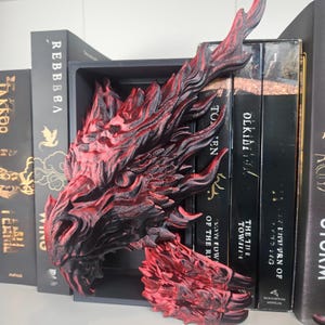 May include: A red and black dragon head bookend, 3D printed, positioned between black books on a shelf. The dragon's detailed design includes textured scales and a fierce expression, adding a fantasy element to the bookshelf.