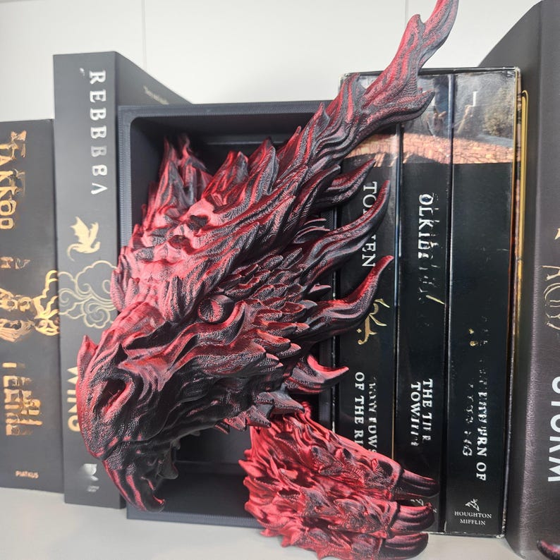 May include: A red and black dragon head bookend, 3D printed, positioned between black books on a shelf. The dragon's detailed design includes textured scales and a fierce expression, adding a fantasy element to the bookshelf.