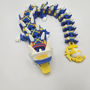 Pode incluir: Uma figura de drag&atilde;o articulada azul e amarela com espinhos brancos. O drag&atilde;o usa um chap&eacute;u branco com "USMC" impresso. Um emblema amarelo e branco est&aacute; preso &agrave; ponta da cauda do drag&atilde;o.