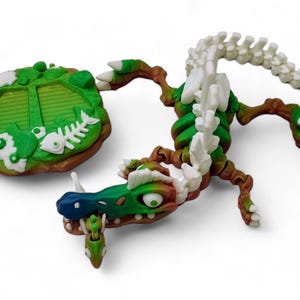 May include: A green and white skeletal dragon figurine with a green and brown base. The dragon has a long tail and a large head with a toothy grin. The base has a green and brown design with white flowers and fish bones.