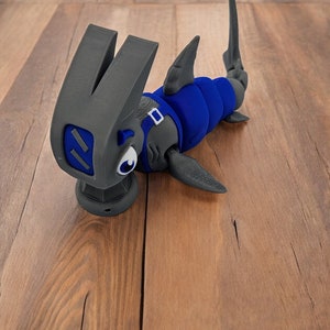 May include: A 3D printed hammerhead shark toy with a blue body and gray fins. The toy is lying on a wooden surface.