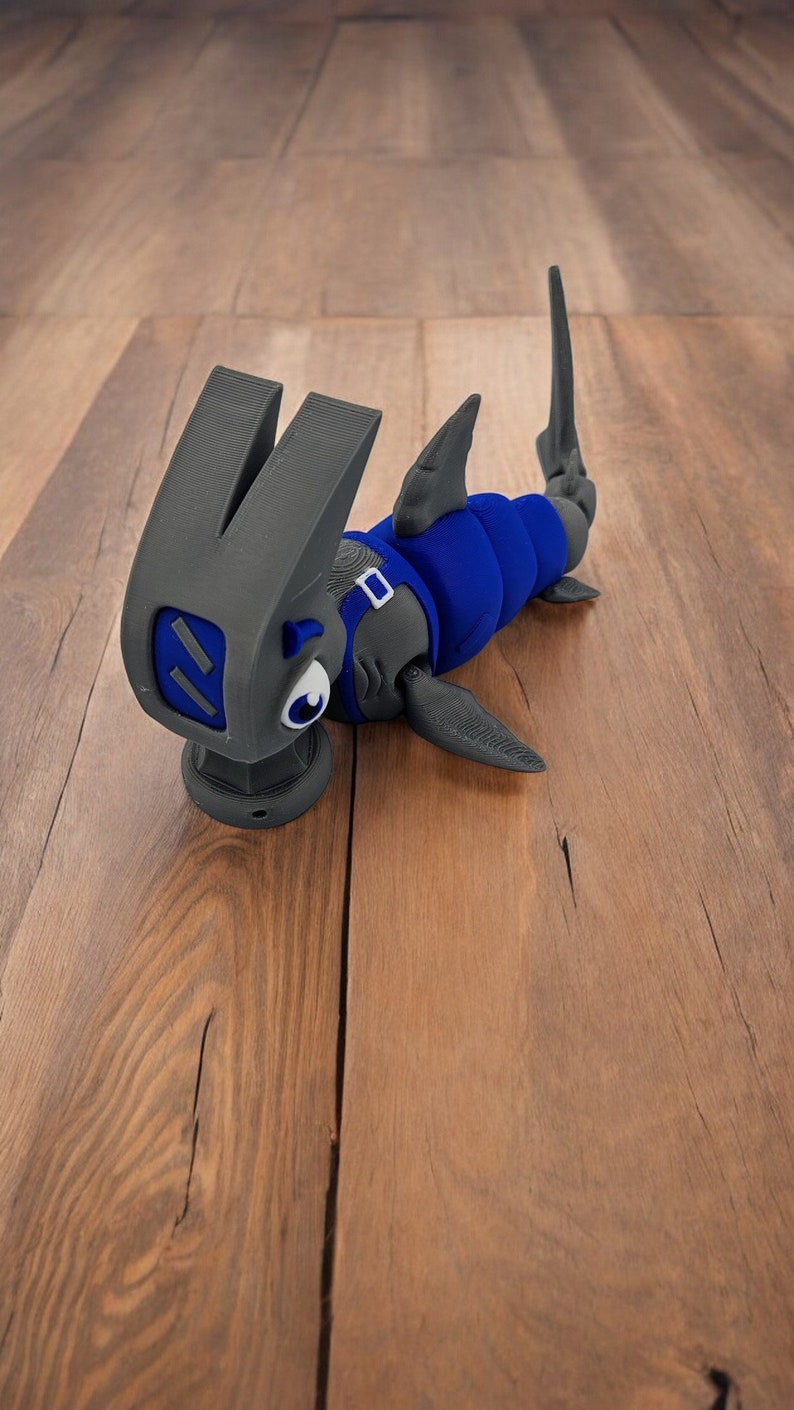May include: A 3D printed hammerhead shark toy with a blue body and gray fins. The toy is lying on a wooden surface.