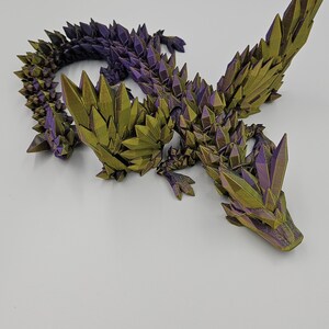 May include: A 3D printed dragon figurine with a green and purple iridescent finish. The dragon is posed in a crawling position with its wings spread out.