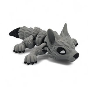 Peut inclure: Une figurine de loup articul&eacute;e grise avec une queue &agrave; bout noir. Le loup pr&eacute;sente des d&eacute;tails, notamment des oreilles pointues, des pattes et un corps textur&eacute;. Les yeux sont noirs avec des reflets blancs. La figurine est couch&eacute;e.