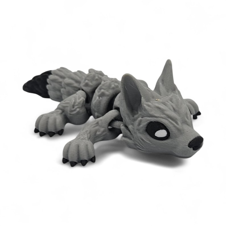 Peut inclure: Une figurine de loup articul&eacute;e grise avec une queue &agrave; bout noir. Le loup pr&eacute;sente des d&eacute;tails, notamment des oreilles pointues, des pattes et un corps textur&eacute;. Les yeux sont noirs avec des reflets blancs. La figurine est couch&eacute;e.