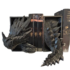 Op de afbeelding: Zwarte boekensteunen met drakenmotief. De boekensteunen zijn ontworpen om op de kop en staart van een draak te lijken, met ingewikkelde details. De kop van de draak bevindt zich links en de staart rechts. Boeken worden tussen de boekensteunen geplaatst.