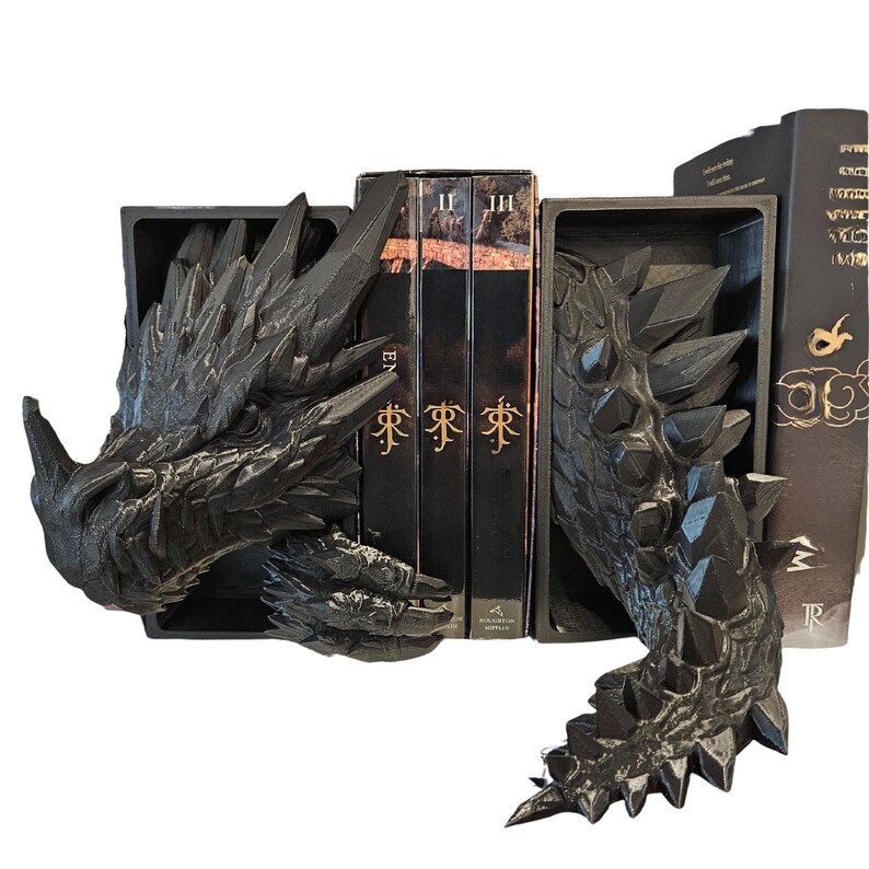Op de afbeelding: Zwarte boekensteunen met drakenmotief. De boekensteunen zijn ontworpen om op de kop en staart van een draak te lijken, met ingewikkelde details. De kop van de draak bevindt zich links en de staart rechts. Boeken worden tussen de boekensteunen geplaatst.