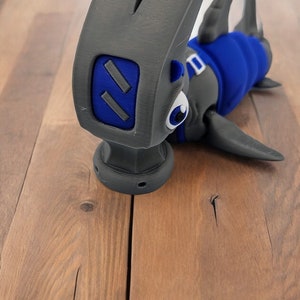May include: A gray hammer with a blue shark head and a white eye. The hammer is on a wooden surface.