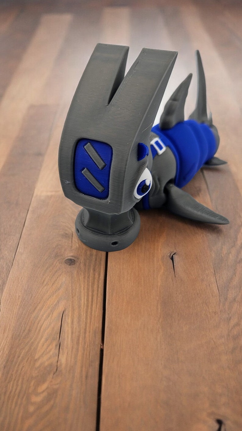 May include: A gray hammer with a blue shark head and a white eye. The hammer is on a wooden surface.