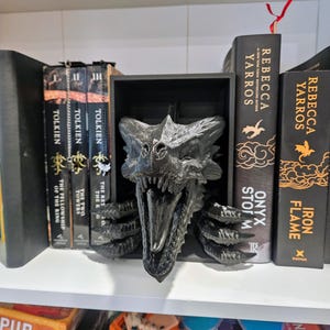 Pode incluir: Um suporte de livros com cabe&ccedil;a de drag&atilde;o preto, com escamas detalhadas e boca aberta. Est&aacute; numa prateleira branca, segurando livros de Tolkien e Rebecca Yarros. A cabe&ccedil;a de drag&atilde;o &eacute; o ponto focal, adicionando um toque de fantasia.