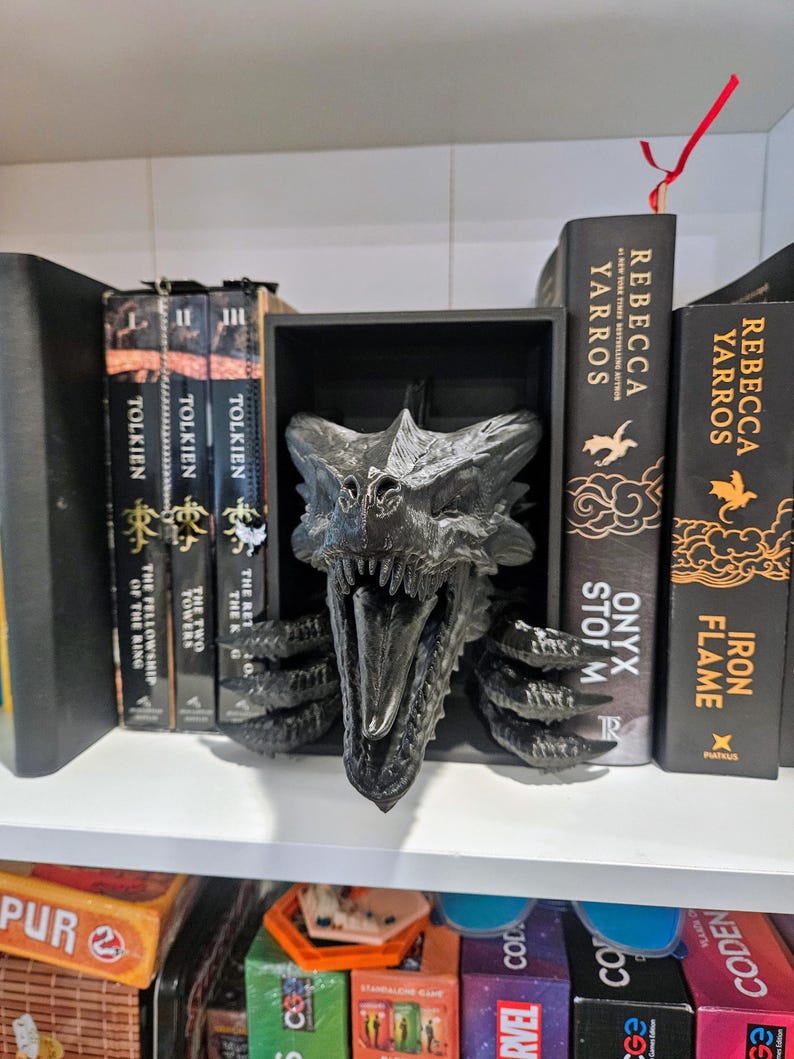 Pode incluir: Um suporte de livros com cabe&ccedil;a de drag&atilde;o preto, com escamas detalhadas e boca aberta. Est&aacute; numa prateleira branca, segurando livros de Tolkien e Rebecca Yarros. A cabe&ccedil;a de drag&atilde;o &eacute; o ponto focal, adicionando um toque de fantasia.