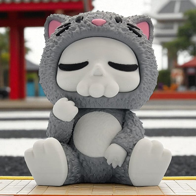 May include: A gray figurine of a cat-like creature with a skull-shaped face, wearing a hooded outfit. The figurine has closed eyes, pink inner ears, and white paws. It is sitting with its legs crossed on a wooden surface.