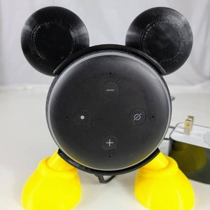 Puede incluir: Un altavoz Amazon Echo Dot de 3.&ordf; generaci&oacute;n negro con un dise&ntilde;o de Mickey Mouse impreso en 3D. El altavoz tiene patas amarillas y orejas negras. El altavoz tiene botones para subir el volumen, bajar el volumen y silenciar.