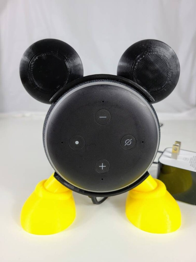 Puede incluir: Un altavoz Amazon Echo Dot de 3.&ordf; generaci&oacute;n negro con un dise&ntilde;o de Mickey Mouse impreso en 3D. El altavoz tiene patas amarillas y orejas negras. El altavoz tiene botones para subir el volumen, bajar el volumen y silenciar.
