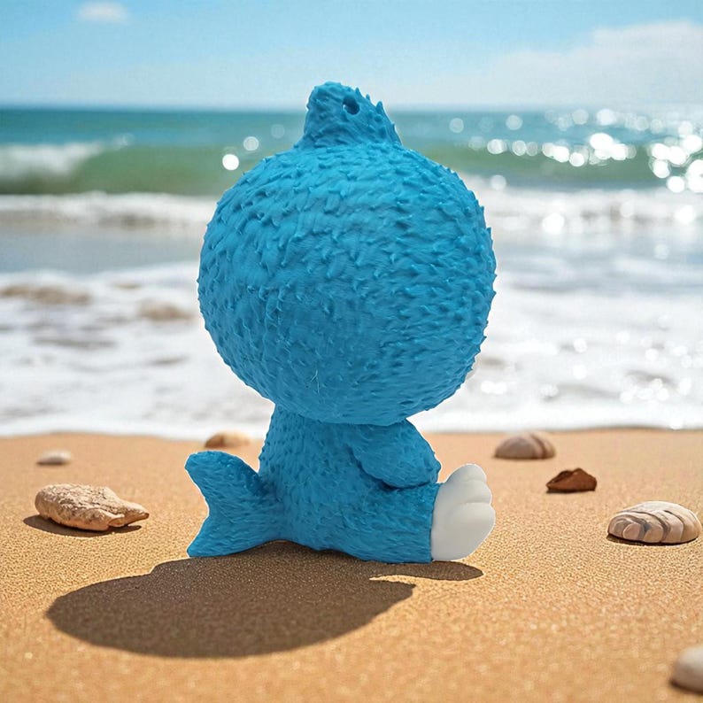 May include: A blue cartoon character with a textured surface sits on a sandy beach, facing the ocean. The figure has a round head, a small tail, and white feet. The background shows the ocean and sky.
