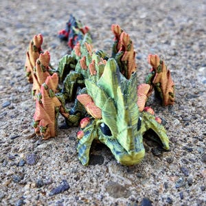 May include: A small, green and orange dragon figurine with spiky wings and a textured surface. The dragon is posed in a crawling position with its head raised.