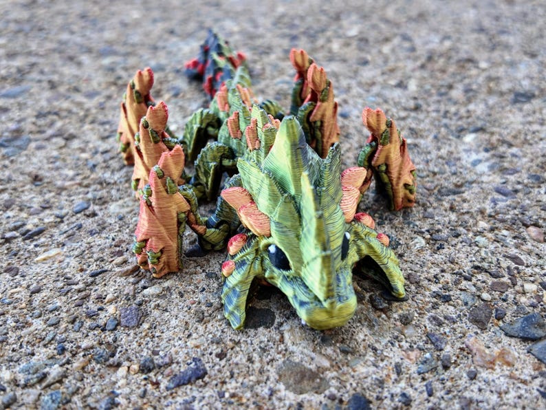 May include: A small, green and orange dragon figurine with spiky wings and a textured surface. The dragon is posed in a crawling position with its head raised.