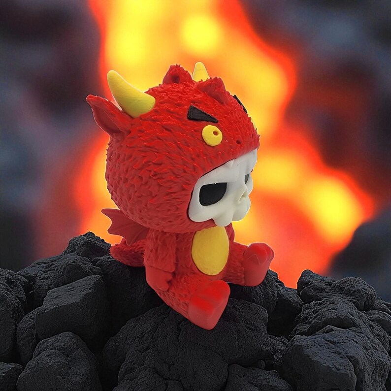 Puede incluir: Un juguete de peluche rojo con un disfraz de diablo con cuernos amarillos, una barriga amarilla y una cara de calavera. El juguete est&aacute; sentado en una pila de rocas de color gris oscuro, con un fondo naranja y amarillo llameante.