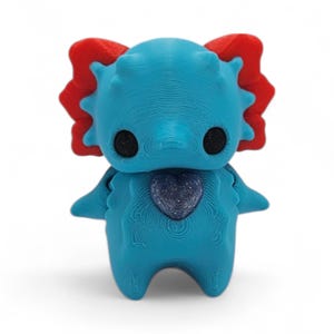 May include: A 3D-printed figurine of a cartoonish creature in shades of blue and red. The figure has large black eyes, a heart-shaped design on its chest, and a textured surface. The creature has a whimsical design.