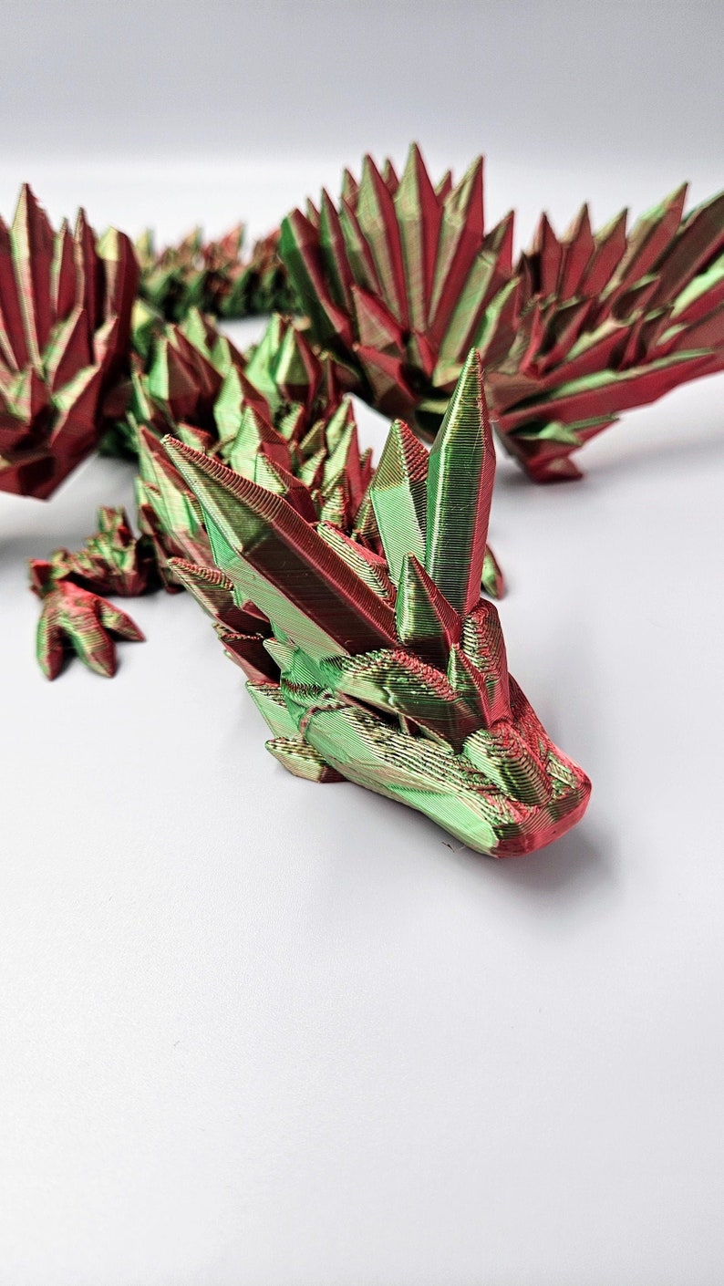 May include: A 3D printed dragon figurine with a green and red iridescent finish. The dragon is posed with its head raised and its wings spread out.