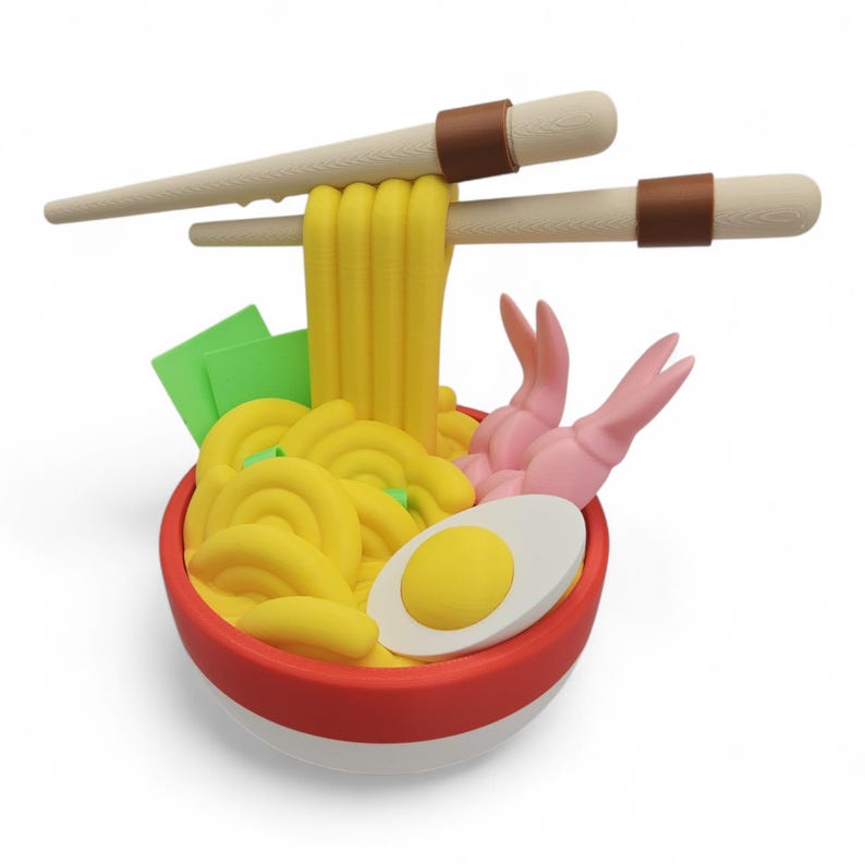 K&ouml;nnte beinhalten: 3D-Darstellung einer Sch&uuml;ssel Ramen mit Essst&auml;bchen. Die rot-wei&szlig;e Sch&uuml;ssel ist mit gelben Nudeln, einem gekochten Ei, rosa Garnelen und gr&uuml;nem Seetang gef&uuml;llt. Beige Essst&auml;bchen halten einige Nudeln hoch.