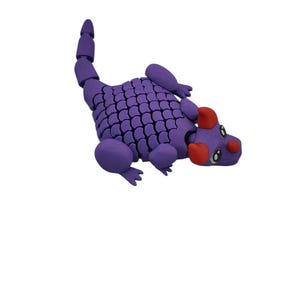 Puede incluir: Un juguete de drag&oacute;n articulado morado con un cuerpo y cola segmentados. El drag&oacute;n tiene detalles rojos en la cabeza y est&aacute; posado sobre un fondo blanco. El juguete est&aacute; dise&ntilde;ado con un patr&oacute;n de escamas.