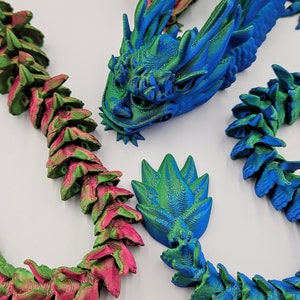 May include: 3D-printed articulated dragons in vibrant colors. The image features a blue and green dragon head, a pink and green dragon, and a blue and green dragon tail. The dragons are made of plastic and have a detailed, textured design.