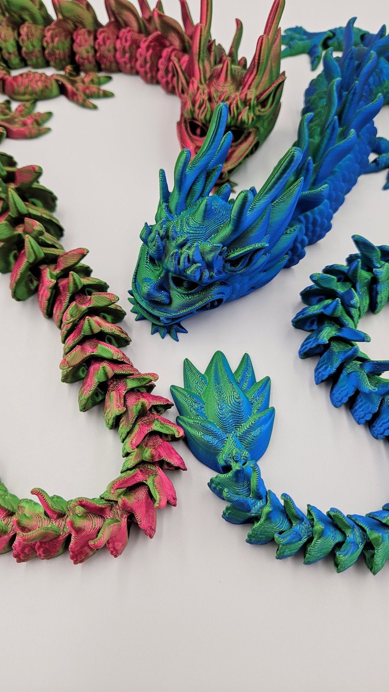 May include: 3D-printed articulated dragons in vibrant colors. The image features a blue and green dragon head, a pink and green dragon, and a blue and green dragon tail. The dragons are made of plastic and have a detailed, textured design.