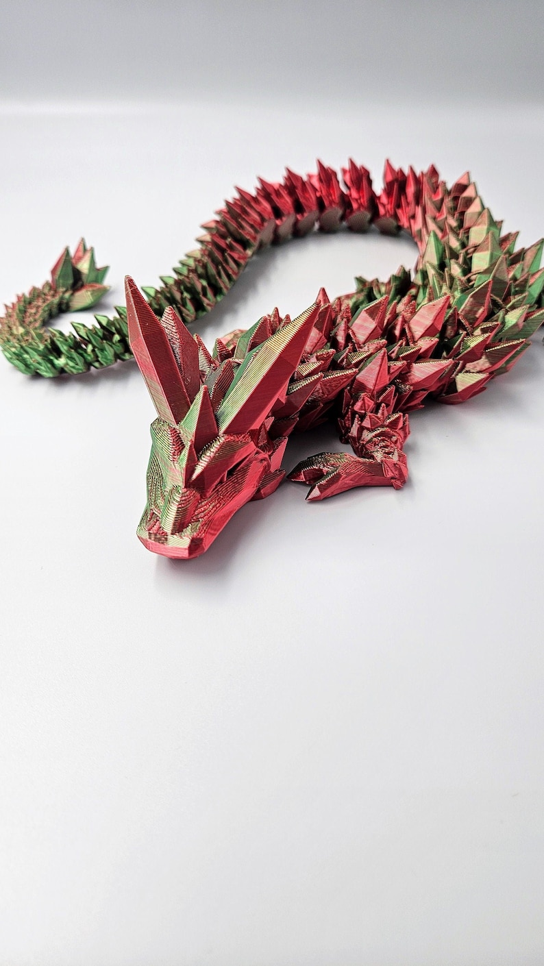 May include: A 3D printed dragon figurine with a red and green iridescent finish. The dragon is curled up in a sleeping position with its head resting on its front paws.