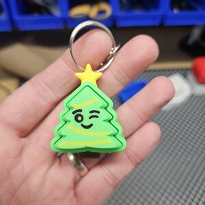 Pode incluir: Um chaveiro de &aacute;rvore de Natal verde e amarelo com uma cara piscando. O chaveiro est&aacute; preso a um chaveiro prateado.