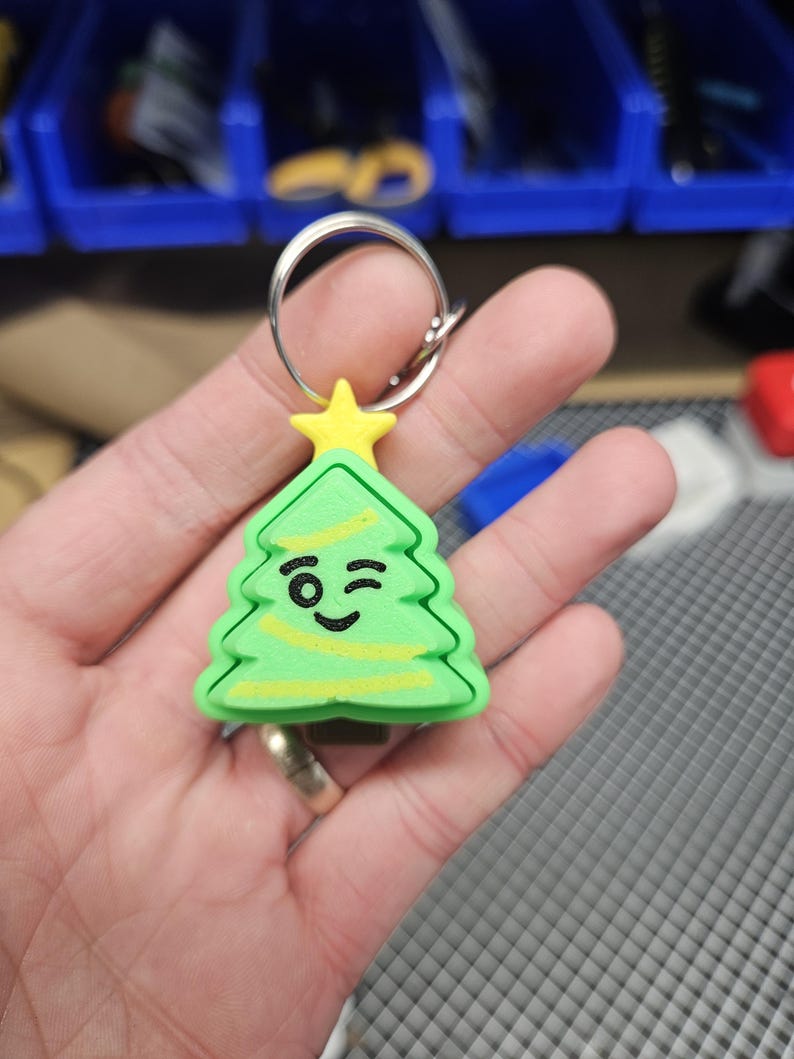 Pode incluir: Um chaveiro de &aacute;rvore de Natal verde e amarelo com uma cara piscando. O chaveiro est&aacute; preso a um chaveiro prateado.