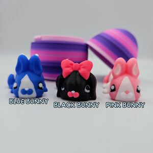 K&ouml;nnte beinhalten: Drei kleine Hasenfiguren, eine blaue, eine schwarze mit einer rosa Schleife und eine rosa. Die Hasen schauen alle nach vorne und haben die Augen geschlossen.