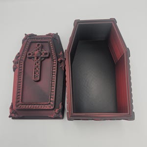 May include: An open, burgundy-colored coffin-shaped box with a detailed cross on the lid. The interior is black, and the box has decorative trim. The coffin is made of a 3D-printed material.