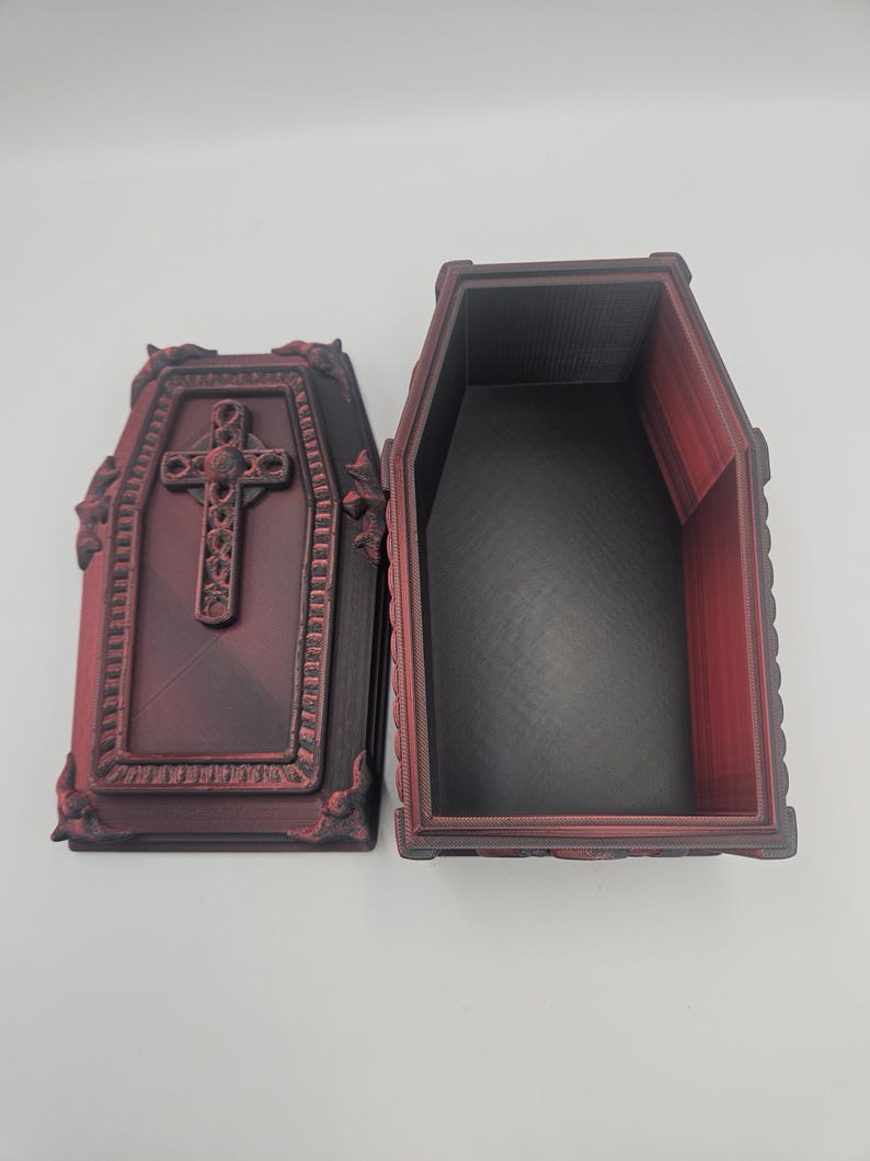 May include: An open, burgundy-colored coffin-shaped box with a detailed cross on the lid. The interior is black, and the box has decorative trim. The coffin is made of a 3D-printed material.