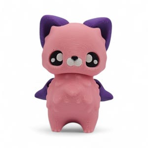 May include: A pink 3D-printed figurine of a stylized animal with large eyes, purple ears, and small purple wings. The figurine has a textured surface and a cute, friendly appearance, perfect for a child's room or as a decorative item.