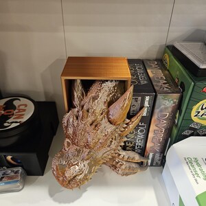May include: A brown dragon head sculpture is displayed on a wooden shelf. The sculpture is made of a textured material and has a detailed design. The shelf is also holding several board games, including "One Night Ultimate Werewolf" and "Daybreak".