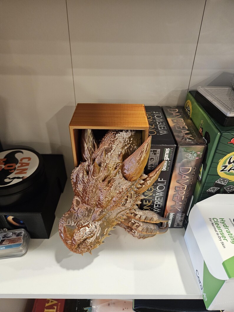 May include: A brown dragon head sculpture is displayed on a wooden shelf. The sculpture is made of a textured material and has a detailed design. The shelf is also holding several board games, including "One Night Ultimate Werewolf" and "Daybreak".