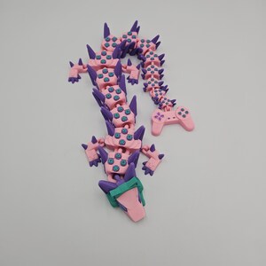 May include: A pink and purple articulated dragon toy with a controller tail. The dragon has a series of connected segments, with purple spikes and teal accents. The controller tail has buttons and a directional pad.