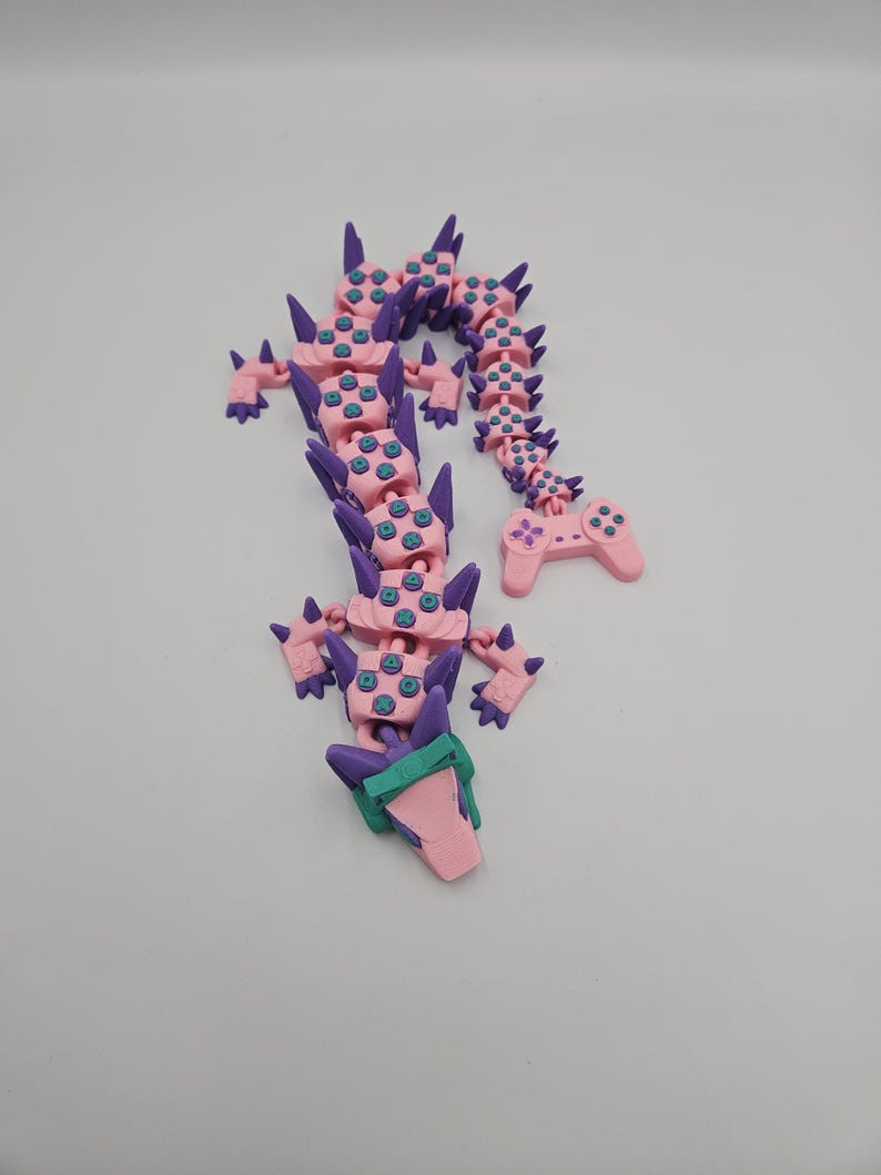 May include: A pink and purple articulated dragon toy with a controller tail. The dragon has a series of connected segments, with purple spikes and teal accents. The controller tail has buttons and a directional pad.