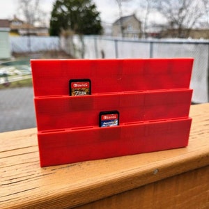 May include: Red plastic Nintendo Switch game card holder with slots for multiple games. Two game cards are visible, one for "The Legend of Zelda: Breath of the Wild" and one for "Super Mario Odyssey".