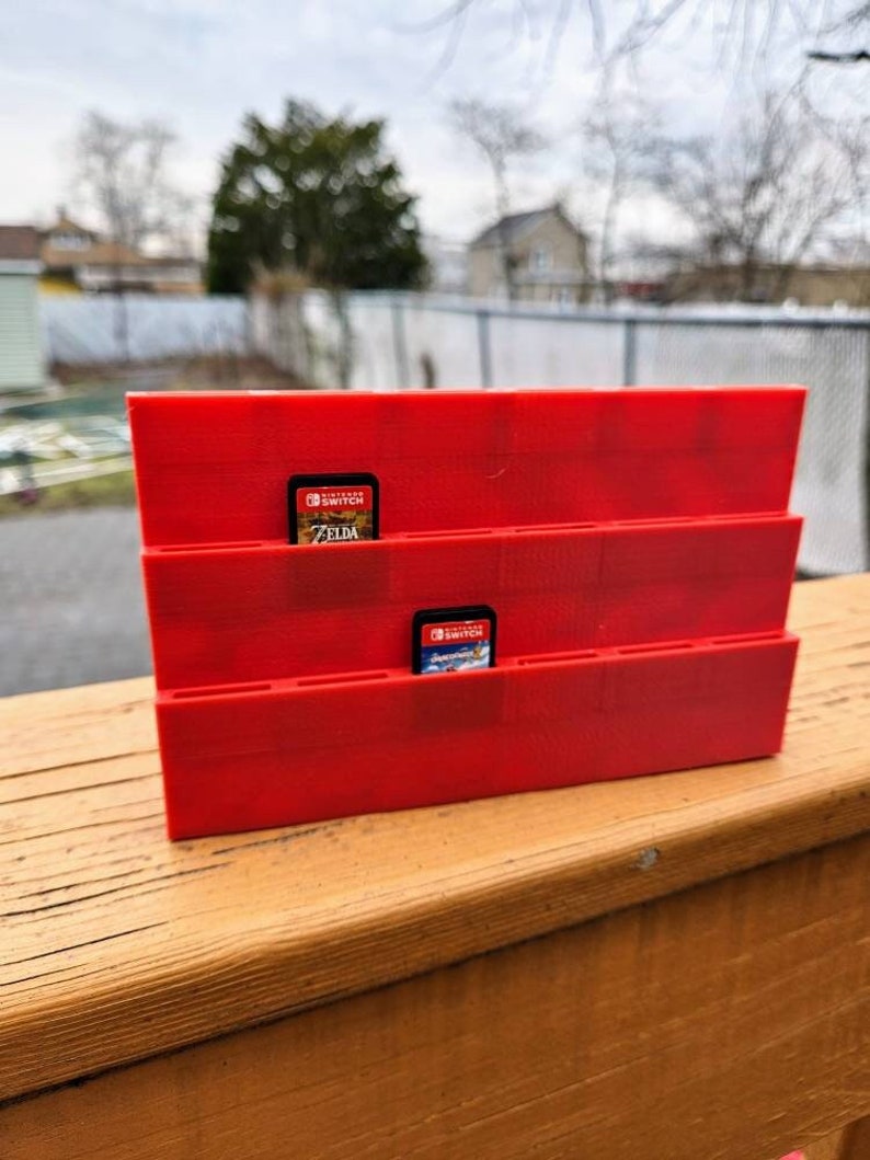 May include: Red plastic Nintendo Switch game card holder with slots for multiple games. Two game cards are visible, one for "The Legend of Zelda: Breath of the Wild" and one for "Super Mario Odyssey".