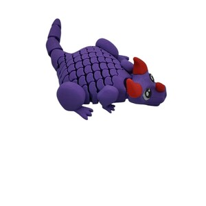 Puede incluir: Un juguete de drag&oacute;n articulado morado con un cuerpo escamado, extremidades redondeadas y una cola segmentada. El drag&oacute;n tiene cuernos rojos y una nariz roja. Los ojos son blancos con pupilas negras. El juguete est&aacute; dise&ntilde;ado para ser posable.