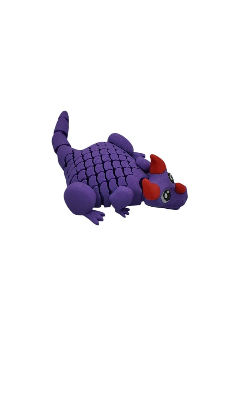 Puede incluir: Un juguete de drag&oacute;n articulado morado con un cuerpo escamado, extremidades redondeadas y una cola segmentada. El drag&oacute;n tiene cuernos rojos y una nariz roja. Los ojos son blancos con pupilas negras. El juguete est&aacute; dise&ntilde;ado para ser posable.