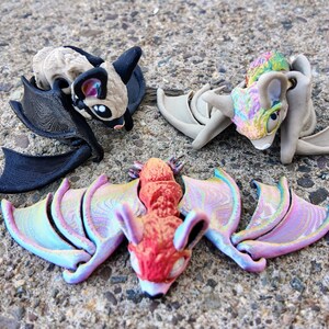 May include: Three small bat figurines with iridescent wings. One bat is black, one is beige with black wings, and one is red with iridescent pink and purple wings.